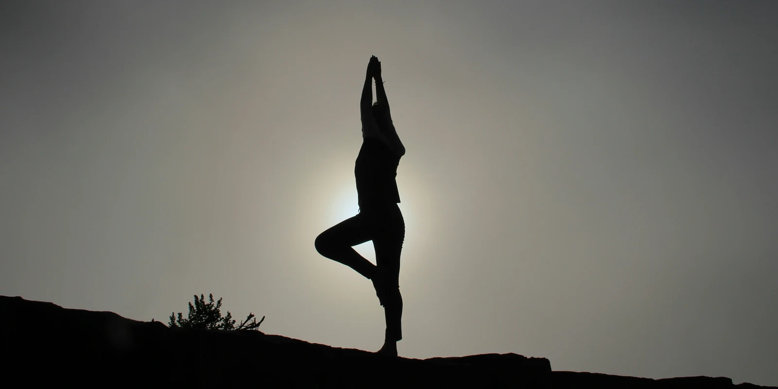 Silhouet van iemand die in de yogahouding 'de boom' op een heuvelrug staat, met de zon vaag achter zich.